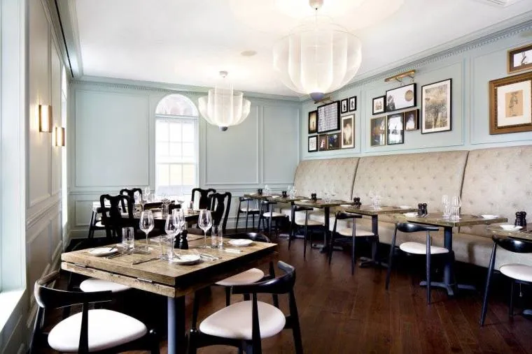 A modern, elegant restaurant interior with light blue walls, wooden floors, and a long upholstered bench. Tables are set with glasses and cutlery, and framed art decorates the walls above the seating.