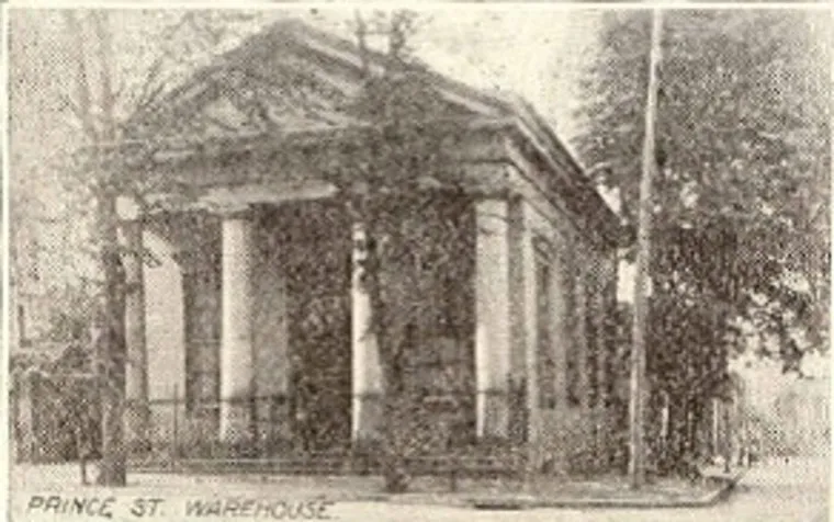 Black-and-white photo of a historic building labeled Prince St. Warehouse, featuring large columns at the entrance and partially obscured by trees.