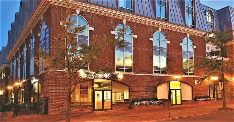 A red brick building with tall arched windows is illuminated in the evening. Trees line the sidewalk, and warm lights from inside and street lamps create a welcoming atmosphere.