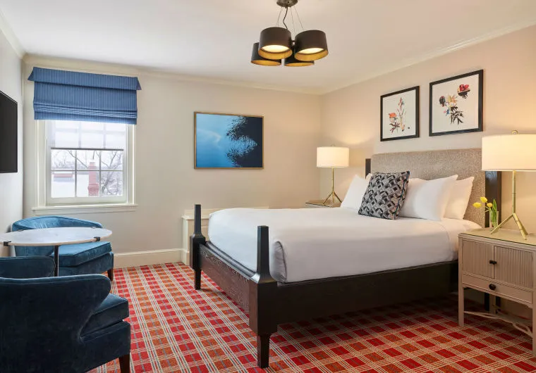 A hotel room with a large bed, two blue armchairs, a small round table, two bedside tables with lamps, wall art above the bed, and a window with a blue shade. The floor is covered with red plaid carpet.