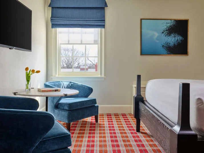 A hotel room with a bed, two blue velvet chairs around a small round table with flowers and a book, a wall-mounted TV, a window with a blue shade, plaid carpet, and a framed abstract artwork on the wall.