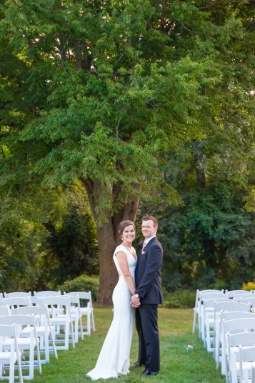 A bride and groom stand together, holding hands and smiling, in an outdoor setting with white chairs arranged on grass and large green trees in the background.