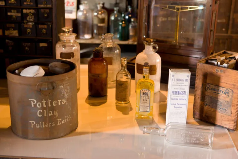 A collection of vintage apothecary bottles, a tin labeled Potters Clay, a glass measuring device, and old packaging displayed on a counter, evoking a historic pharmacy setting.
