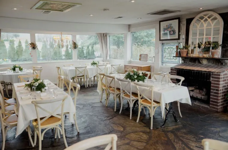 A bright, cozy restaurant dining room with white tablecloths, wooden chairs, floral centerpieces, large windows, and a brick fireplace decorated with plants and framed pictures.
