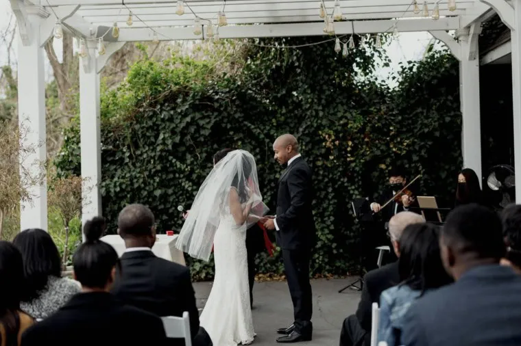 A bride and groom stand facing each other at an outdoor wedding ceremony under a white pergola, surrounded by greenery and guests seated in rows, while musicians play nearby.