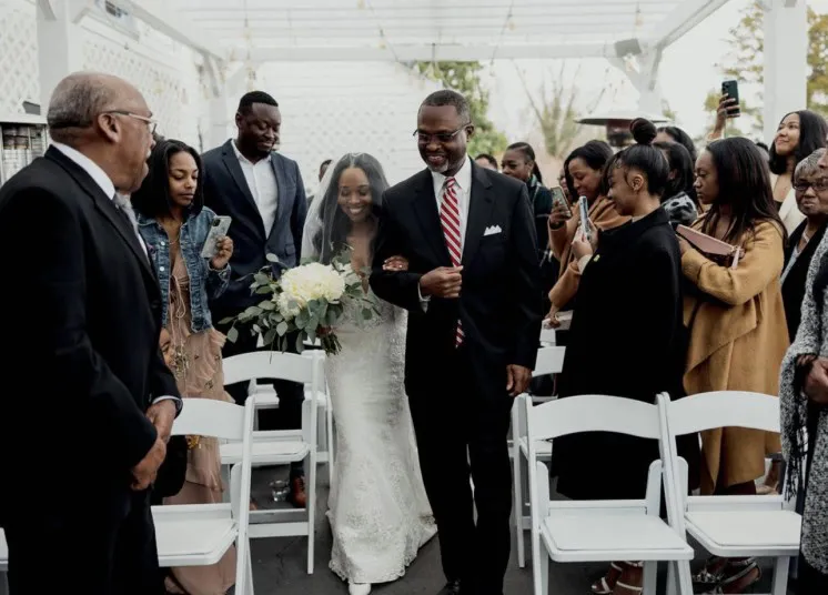 A smiling bride holding a bouquet walks down the aisle arm-in-arm with a man in a black suit and striped tie, surrounded by seated and standing guests taking photos at an outdoor wedding ceremony.