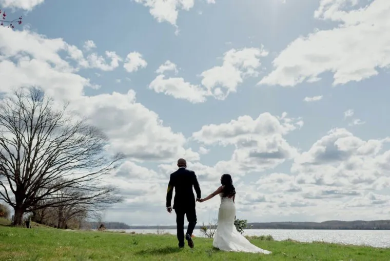 A bride and groom hold hands and walk across a grassy field toward a lake under a partly cloudy sky, with trees and water in the background.