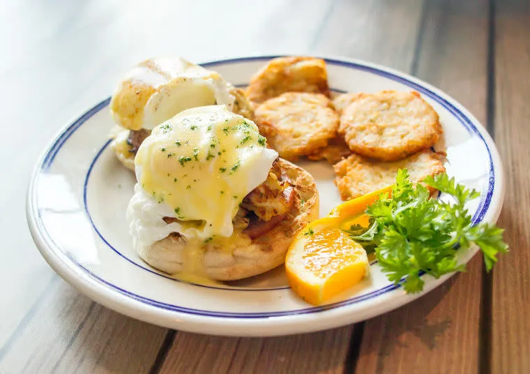 A plate with eggs Benedict topped with hollandaise sauce, served with crispy hash browns, orange slices, and a sprig of parsley on the side.