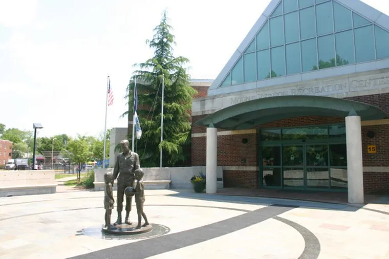 A bronze statue of an adult and two children stands outside a brick building with large glass windows. Two flagpoles with American flags are visible near the entrance and trees surround the area.