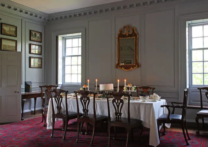 Elegant dining room with a long table set for a meal, white tablecloth, candles, and a cake; wooden chairs surround the table. Large windows, framed art, and a gold mirror decorate the gray paneled walls.
