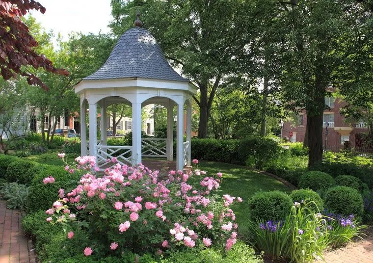 A white gazebo stands in a lush garden surrounded by blooming pink flowers, green bushes, and tall trees, with brick paths and buildings visible in the background.