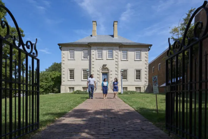 Three people walk on a brick path toward a historic two-story stone house with chimneys, framed by open black iron gates, under a blue sky.