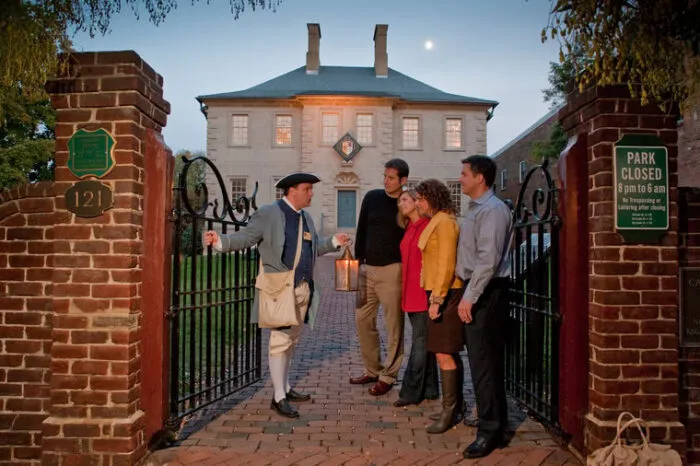 A man in colonial attire holding a lantern welcomes four casually dressed adults at the open gates of a historic brick building at dusk, with a glowing light from the entrance and signs on the gate pillars.