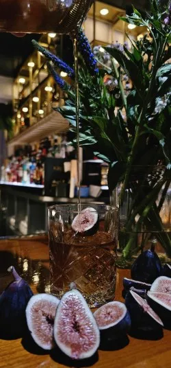 A cocktail is being poured into a glass filled with figs on a bar counter, surrounded by fresh figs, with a vase of flowers and shelves of bottles in the blurred background.