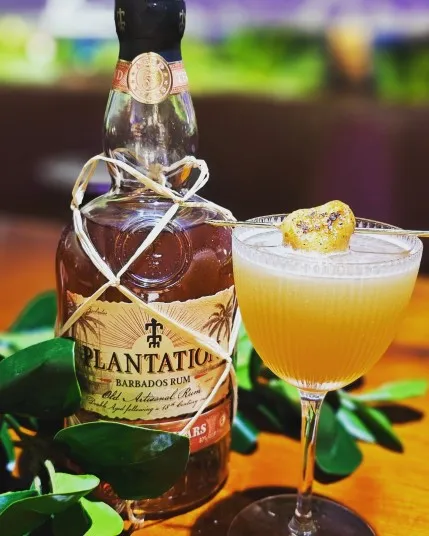 A bottle of Plantation Barbados Rum sits next to a coupe glass filled with a light yellow cocktail, garnished with a caramelized banana slice, on a wooden table with green leaves for decoration.