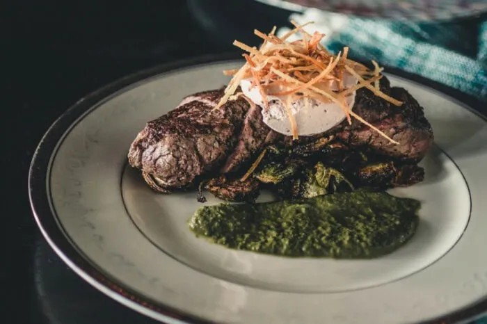 A plated steak topped with a poached egg and crispy shredded garnish, served with roasted vegetables and a green sauce on a white, rimmed plate.