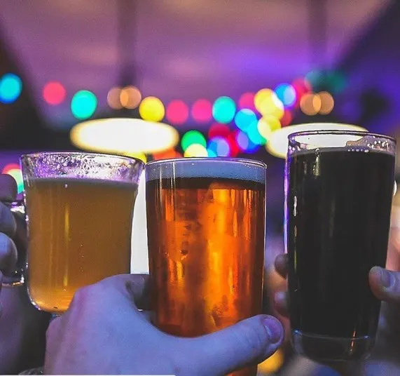 Three hands holding different types of beer in mugs and glasses, raised together in a toast. The background is blurred with colorful, festive lights creating a lively atmosphere.