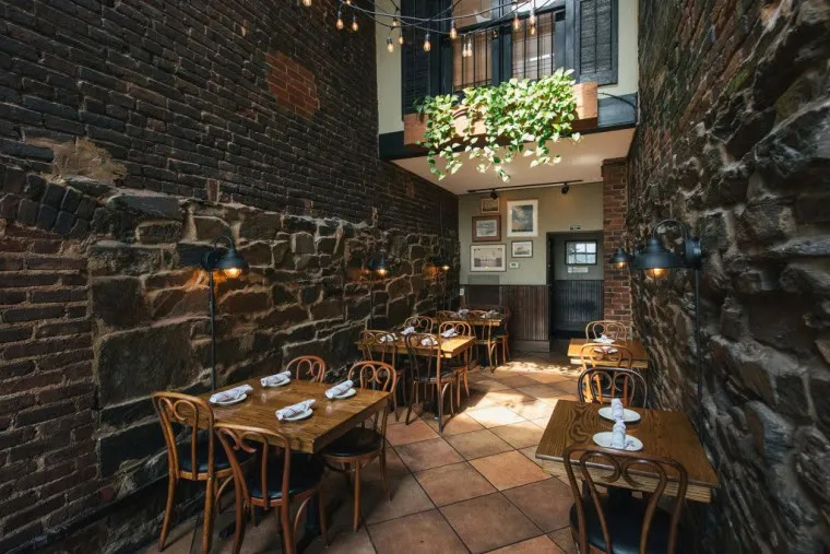 Cozy restaurant with exposed brick and stone walls, wooden tables and chairs, napkins neatly placed, string lights above, framed art on the wall, and a balcony with green plants letting in natural light.