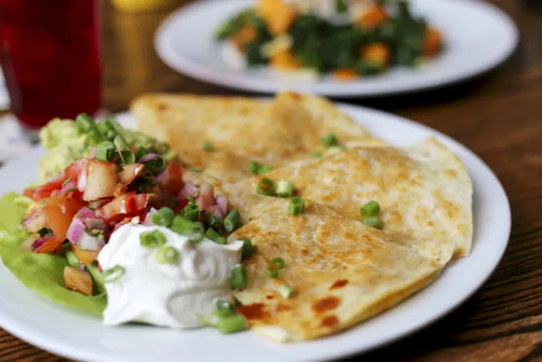 A plate with golden quesadillas, topped with chopped green onions, served with sour cream, guacamole, and pico de gallo; another plate of food is blurred in the background.