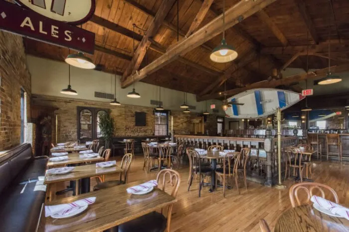 Spacious restaurant interior with wooden tables and chairs, exposed brick walls, high wooden ceiling, and large windows. Tables are set with napkins. A vintage sign reading ALES hangs from above.