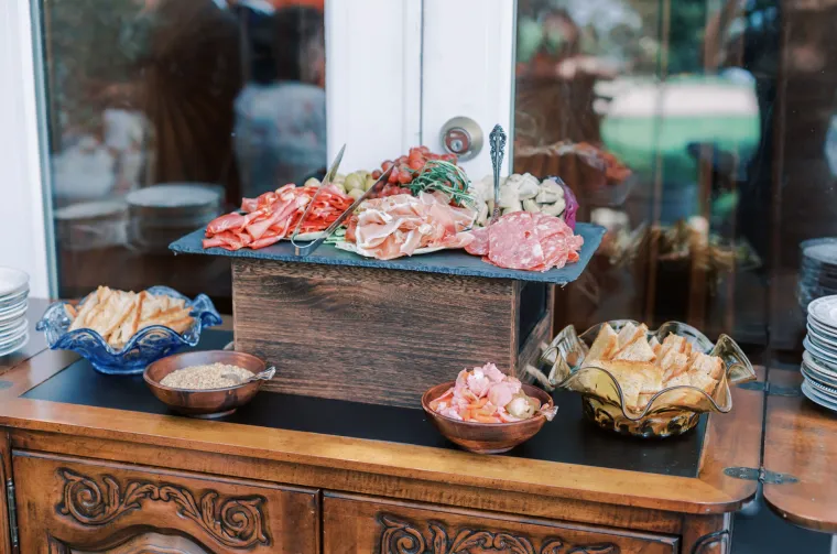 A wooden table holds a charcuterie board with assorted cured meats, cheese, olives, and greens, surrounded by bowls of bread slices, dips, and spreads; stacked plates sit to the side, ready for serving.