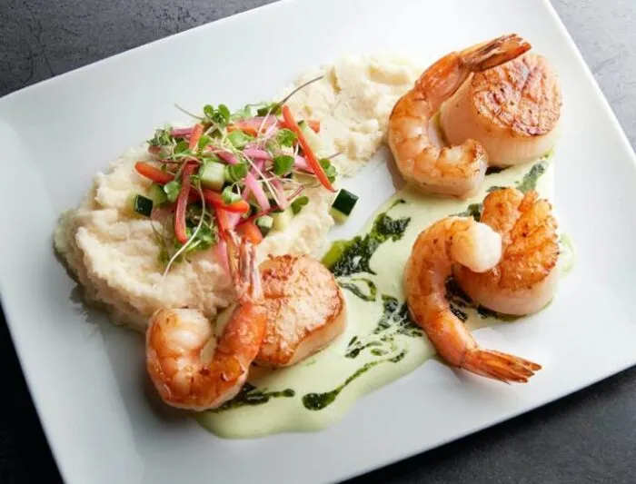 A white plate with seared scallops and shrimp on a green herb sauce, served with mashed potatoes topped with julienned vegetables and microgreens.