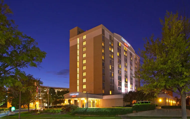 A mid-rise Courtyard by Marriott hotel is illuminated at night, with warm lights glowing from its windows. Trees and bushes frame the building, and the sky is deep blue, indicating dusk or early evening.