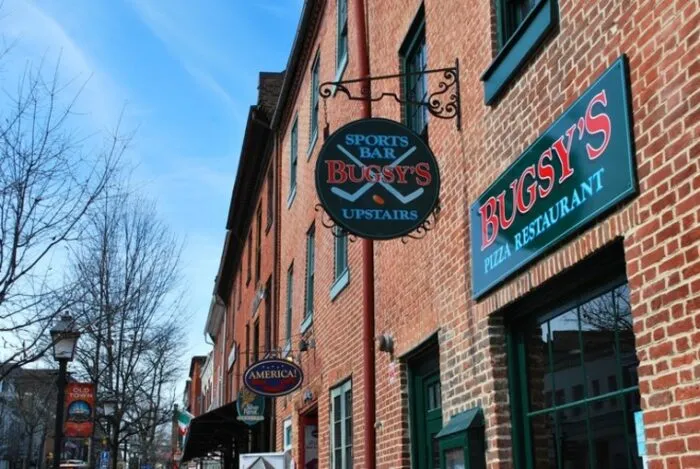 A brick building with signs for Bugsys Sports Bar Upstairs and Bugsys Pizza Restaurant above a sidewalk; leafless trees and a blue sky are in the background.