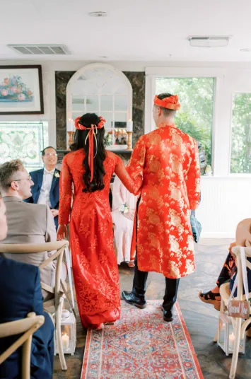 A couple dressed in traditional red Vietnamese attire walks arm-in-arm down the aisle at an indoor wedding ceremony, with seated guests watching and natural light streaming through the windows.