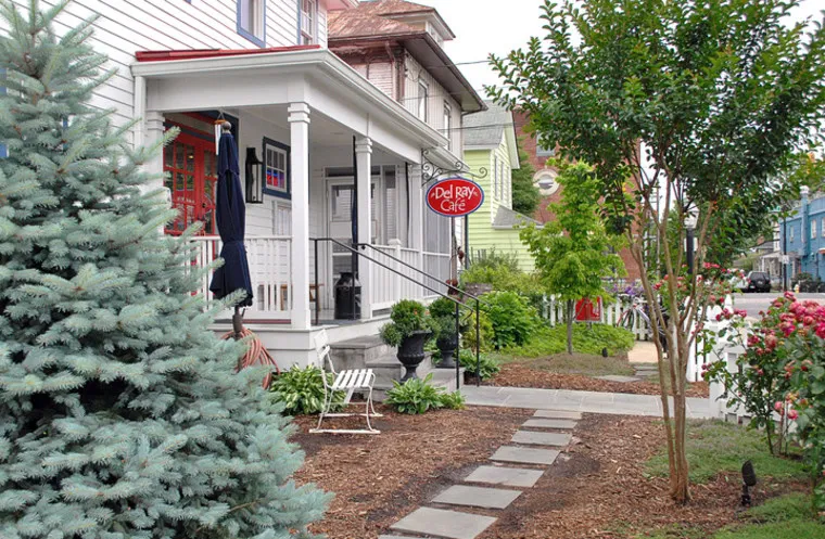 A quaint café with a red Del Ray Café sign hangs beside a white porch, surrounded by green trees, shrubs, and a stone path leading up to the entrance in a suburban neighborhood.