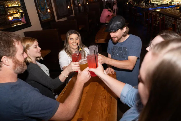 Five people sitting around a wooden table in a bar, raising their drinks together in a toast and smiling. The atmosphere is lively and casual, with dim lighting and booths in the background.