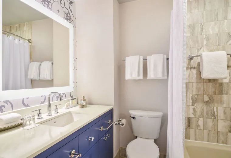 Modern bathroom with a blue vanity, large mirror, white countertop, toilet, towel rack with white towels, and a bathtub with beige tiles and a white shower curtain.