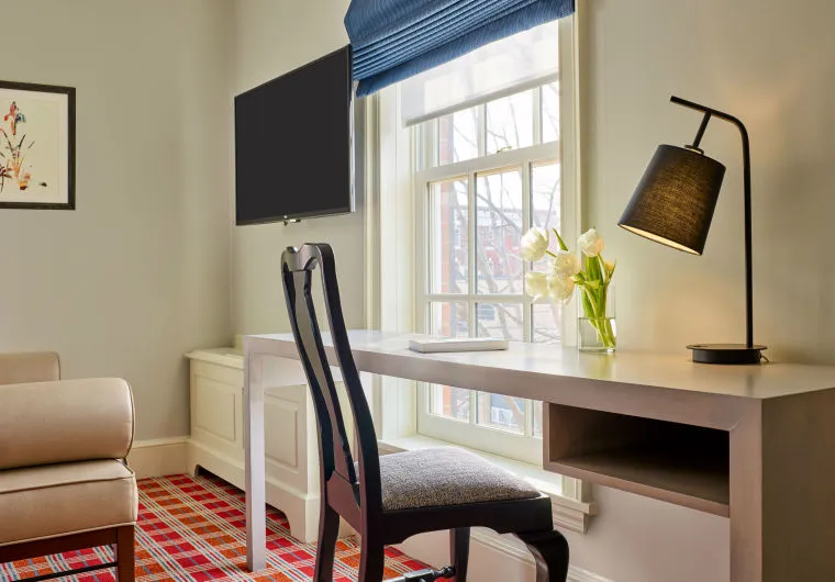 A modern home office with a white desk, black chair, desk lamp, vase of white tulips, and a tablet. Theres a large window with blue blinds, a wall-mounted TV, and red plaid carpet. An armchair and framed artwork are also visible.
