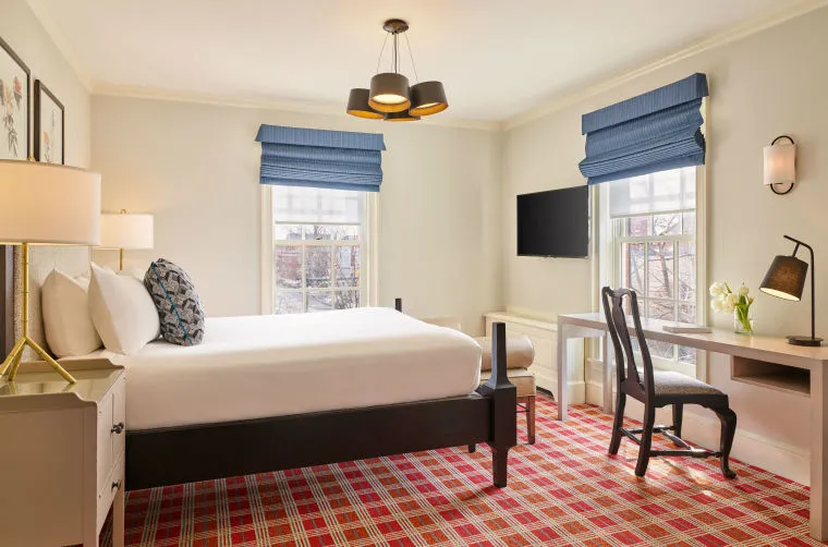 A bright hotel room with a large bed, blue window shades, red plaid carpet, a desk with a chair, a wall-mounted TV, and modern lighting fixtures above. Two windows let in natural light.