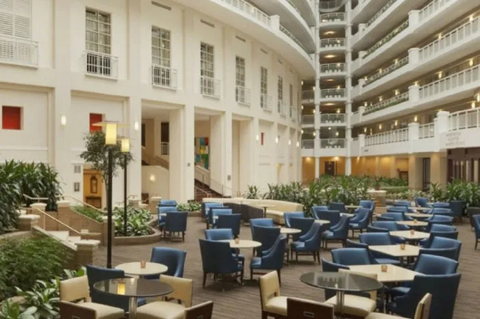 A spacious hotel atrium with tall white walls, multiple floors of balconies, blue and beige chairs around round tables, potted plants, and warm lighting creating a welcoming atmosphere.