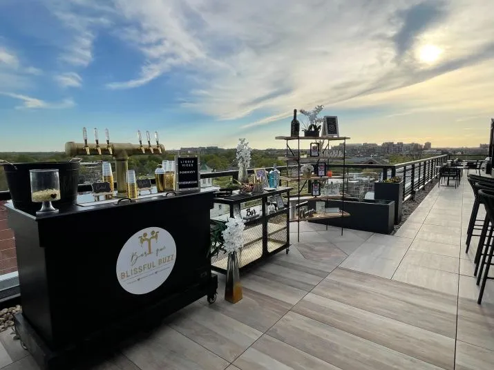 Outdoor rooftop bar setup at sunset with a Blissful Buzz drink cart, champagne bottles, glasses, and city views in the background. Tall tables and chairs line the tiled patio.