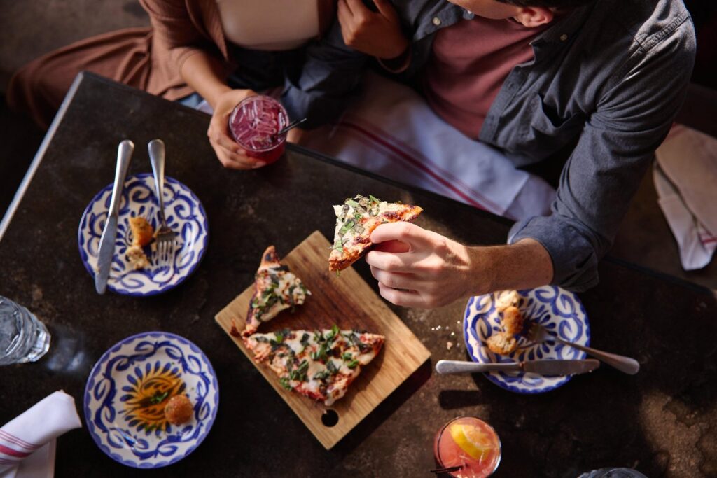 A couple sits at a table with patterned plates, drinks, and a wooden board holding pizza slices. One person holds up a slice, and their drinks are garnished with fruit. The scene looks casual and inviting.
