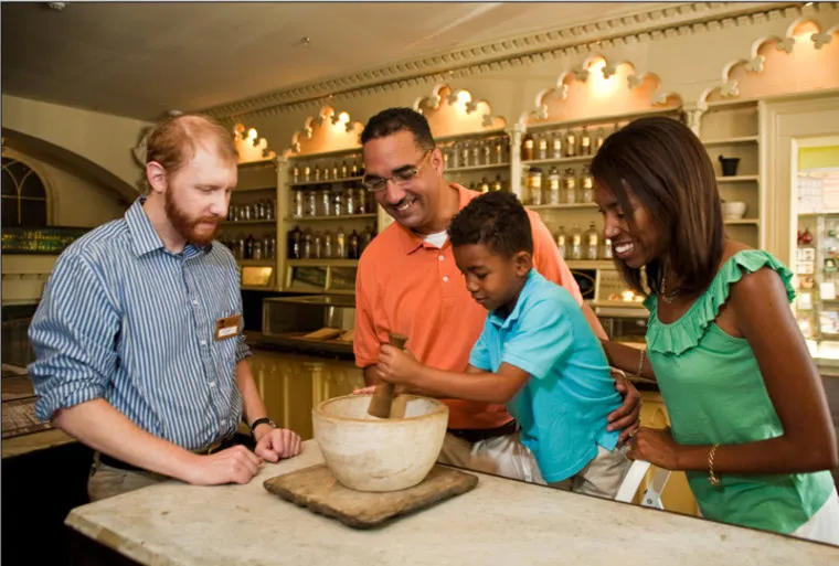 A young boy grinds something in a large mortar and pestle, watched by two adults and a museum staff member behind a counter lined with antique jars and shelves. Everyone appears engaged and smiling.