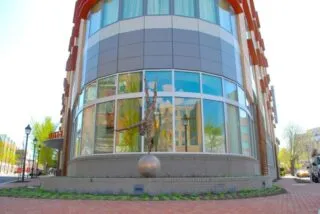 A sculpture of a person balancing upside down on a sphere is displayed in front of a modern, curved glass building with brick pavement and a street in the background.