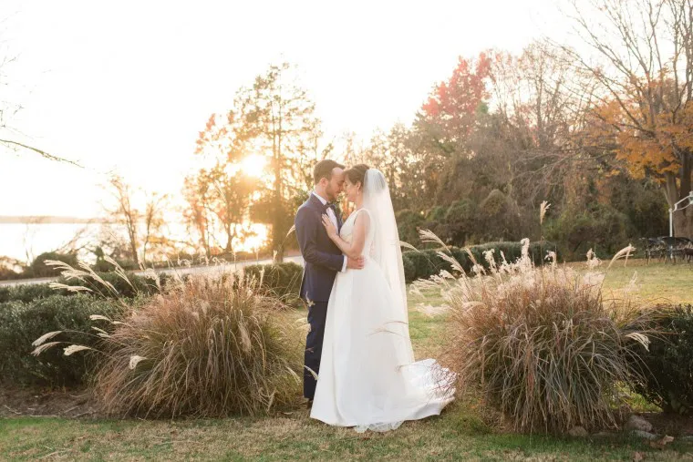 A bride and groom stand facing each other and embrace outdoors at sunset, surrounded by tall grasses and trees with autumn foliage, creating a warm, romantic atmosphere.