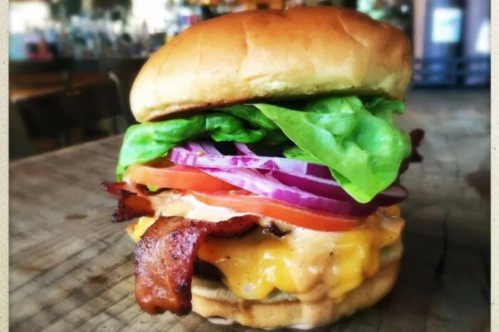 A close-up of a cheeseburger with crispy bacon, cheddar cheese, lettuce, tomato, red onion, and sauce in a toasted bun, set on a wooden surface with a blurred background.