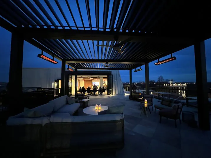 A modern rooftop lounge at night, softly lit by candles and overhead heaters, with cushioned seating and tables. The interior, visible through large windows, glows warmly as people dine inside.