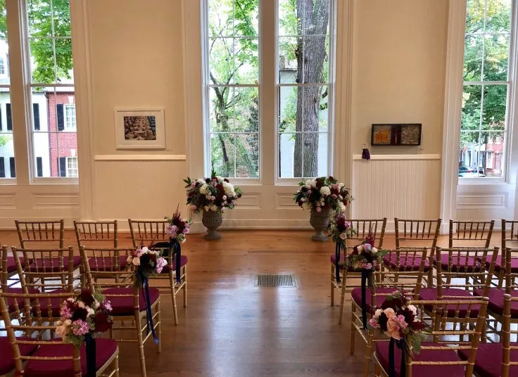 A bright wedding ceremony room with rows of gold chairs with purple cushions and floral decorations, facing large windows and two flower arrangements at the front. Artwork is on the walls and trees are visible outside.