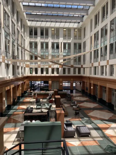 A spacious, sunlit atrium with high ceilings, large windows, and modern lounge seating arranged on a patterned tile floor. Natural light fills the area through a glass roof above.