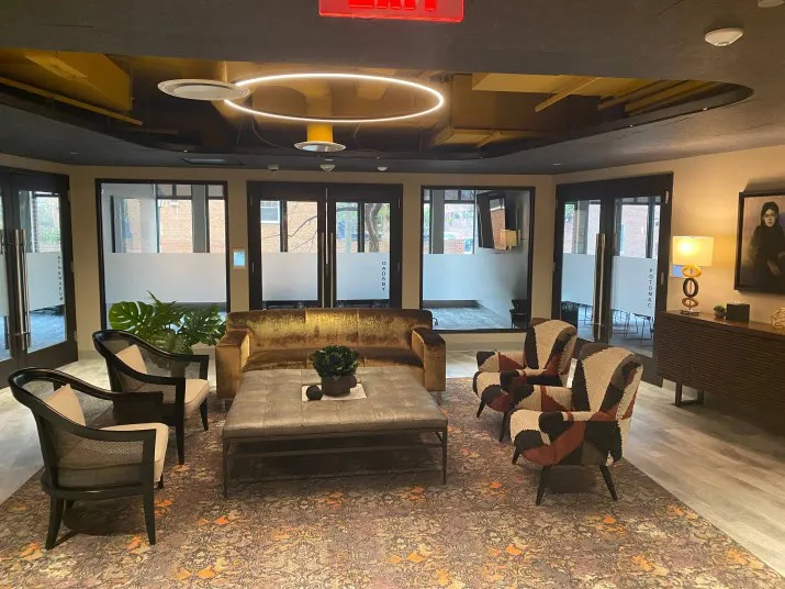 A modern lobby with glass doors, a gold velvet sofa, patterned armchairs, a large ottoman, potted plants, and a circular light fixture on the ceiling. Warm lighting and a rug create a welcoming atmosphere.