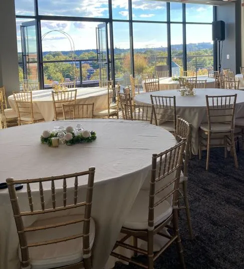 Round tables with white tablecloths and simple floral centerpieces are arranged in a bright event room with open windows, gold chairs, and a scenic view of trees and sky outside.