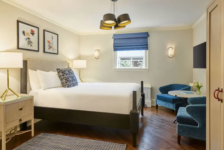 A bright, modern hotel room with a large neatly made bed, two bedside tables with lamps, blue upholstered chairs around a small table, and a window with a blue Roman shade. Artwork decorates the walls.