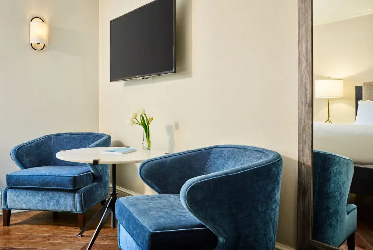 Two blue velvet chairs and a round white table with a small vase of flowers are positioned near a wall-mounted TV and a tall mirror in a bright, modern hotel room.