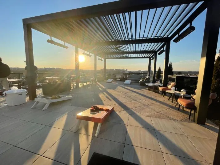 A rooftop patio at sunset with lounge chairs, tables, heaters, and a cornhole game set under a pergola. Long shadows stretch across the tiled floor, creating a warm, inviting atmosphere.