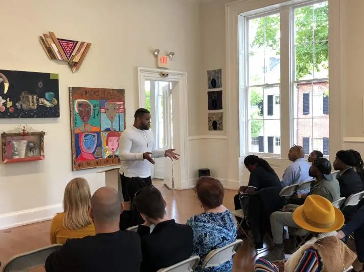 A man stands and speaks to a seated audience in an art gallery, with colorful paintings and large windows in the background. The attendees listen attentively.